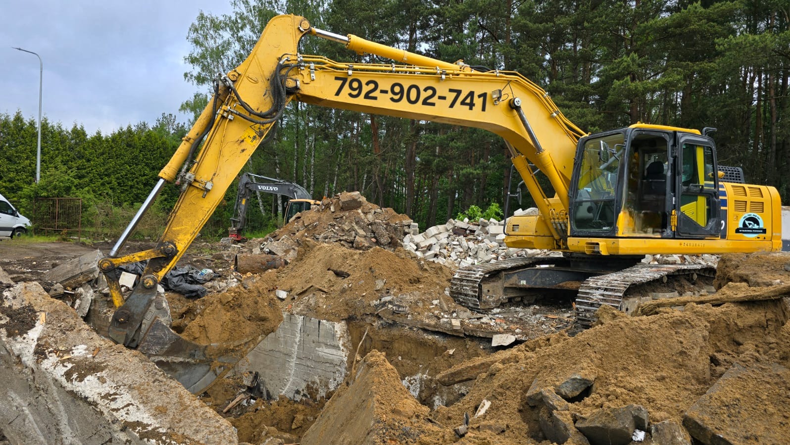 Żółta koparka na gruzowisku z numerem telefonu na ramieniu, w tle zieleń drzew. Widoczny inny sprzęt budowlany i fragment busa.