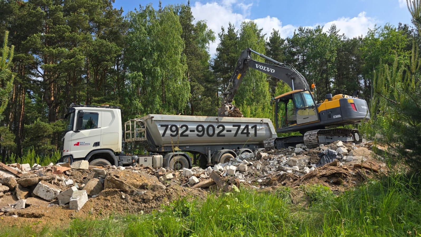 Żółto-szara koparka Volvo ładuje gruz do ciężarówki z numerem telefonu na tle zieleni. Widok na etap prac ziemnych lub wywóz gruzu po rozbiórce.