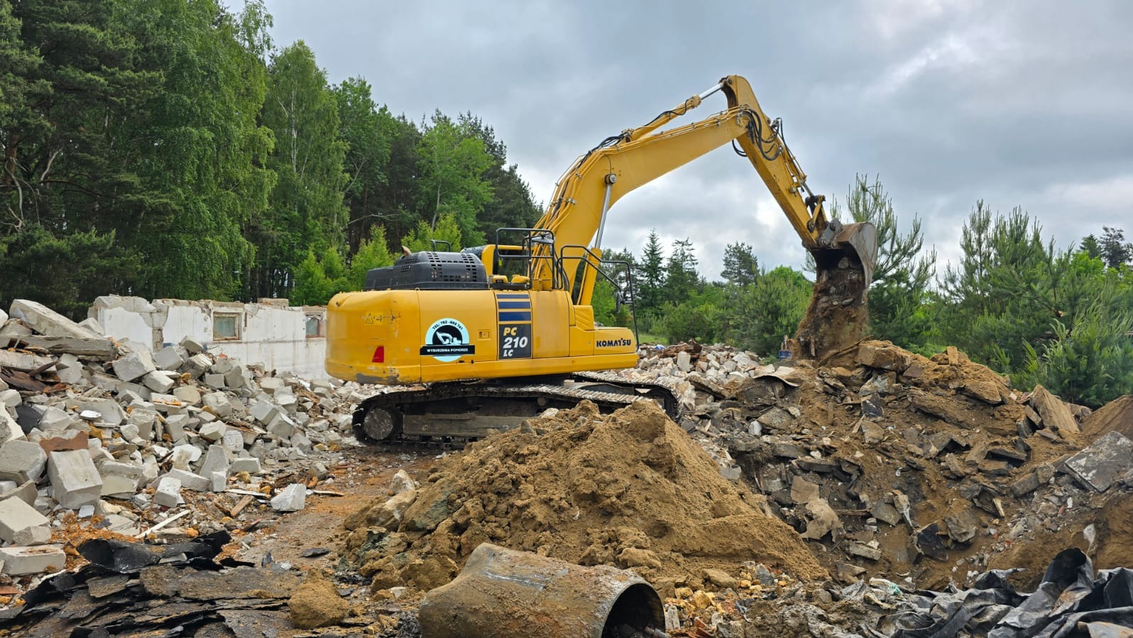 Żółta koparka Komatsu PC210 LC podczas prac rozbiórkowych, z gruzem i elementami budynku w tle, otoczona zielenią drzew. Praca w toku, pochmurne niebo.