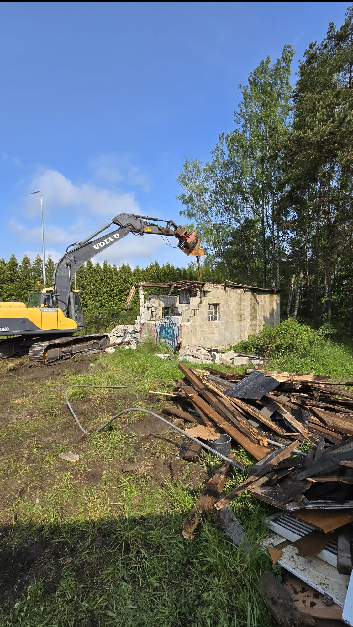 Żółta koparka Volvo z hydraulicznymi nożycami rozbiera murowany budynek. W tle drzewa i błękitne niebo. Fragmenty gruzu i drewna na pierwszym planie.