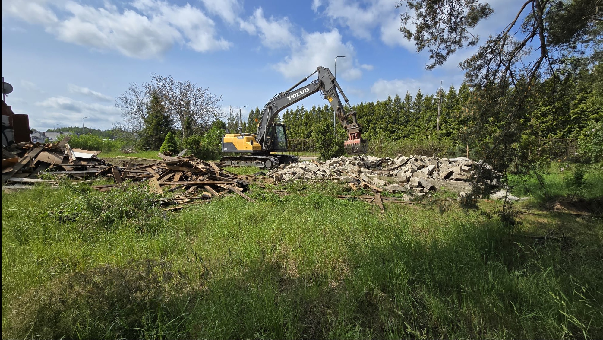 Żółto-szara koparka Volvo z chwytakiem hydraulicznym usuwa gruz i drewno z terenu zielonego, na tle drzew i błękitnego nieba.