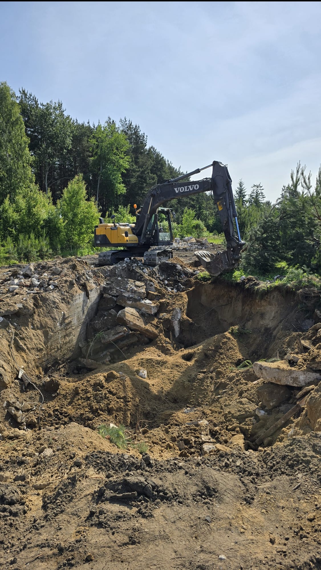 Żółto-czarna koparka Volvo na skraju wykopu z gruzem i ziemią, w tle drzewa, błękitne niebo. Widok z dołu, podkreślający głębokość wykopu i moc maszyny.