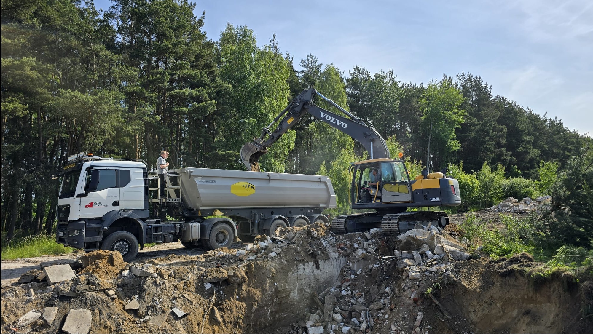 Żółto-czarna koparka Volvo ładuje gruz do szarej ciężarówki MAN Wan-Dróg na tle zieleni drzew. Operator nadzoruje proces.