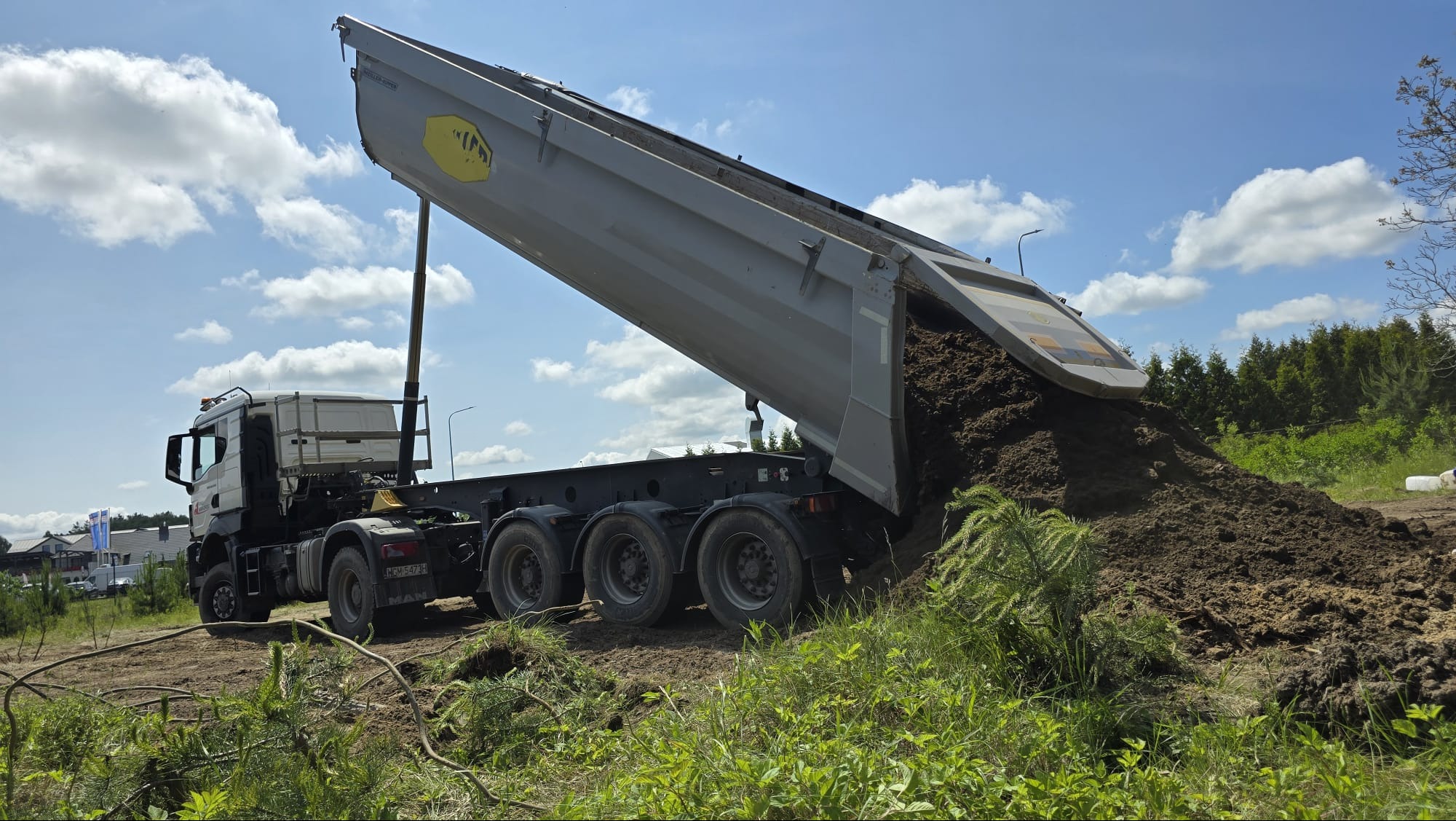 Wyładowywanie ziemi z wywrotki na tle błękitnego nieba z chmurami i zieleni. Widoczny fragment lasu w oddali. Ziemia tworzy mały nasyp.