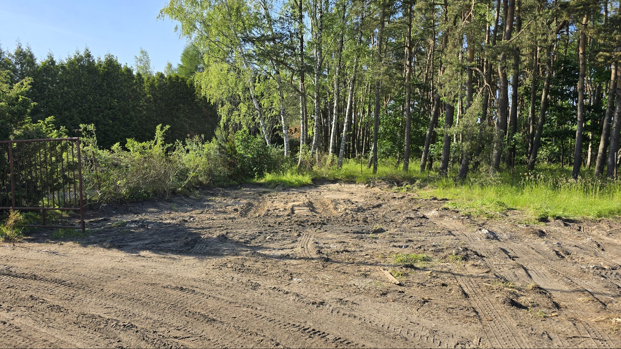 Teren po niwelacji w Chmielnie z widocznymi śladami opon, otoczony zielenią. W tle drzewa i krzewy. Brama wjazdowa po lewej stronie kadru.