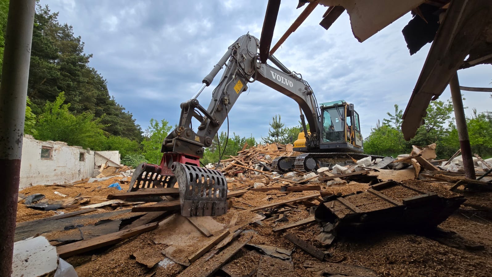 Ciężki sprzęt Volvo w trakcie rozbiórki budynku, z gruzem i elementami konstrukcyjnymi w tle, na tle zieleni i pochmurnego nieba. Ujęcie z dołu.