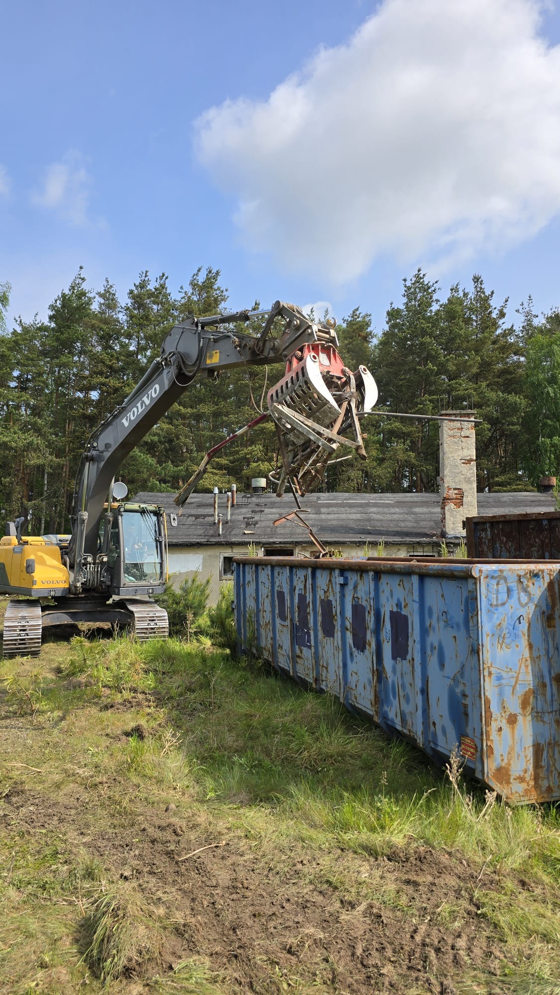 Żółta koparka Volvo z chwytakiem umieszcza metalowe elementy w niebieskim, zardzewiałym kontenerze. W tle budynek i drzewa, błękitne niebo z chmurami.
