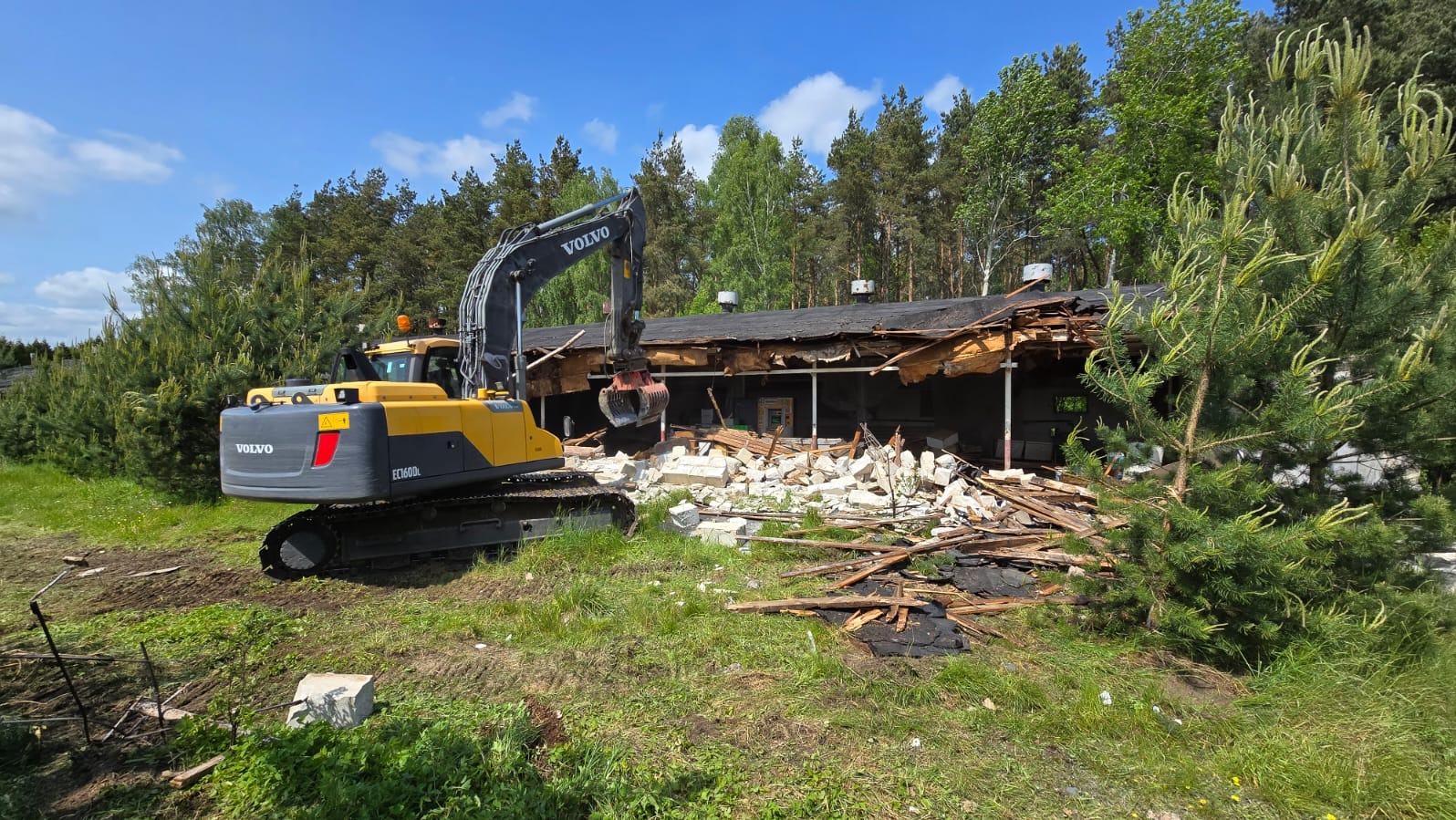 Żółto-szara koparka Volvo rozbiera budynek z gruzem na trawiastym terenie, z drzewami w tle, w słoneczny dzień. Widoczny zniszczony dach i fragmenty ścian.