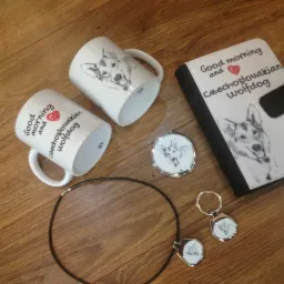 Dwa białe kubki z napisem 'Good morning and Czechoslovakian Wolfdog' i rysunkiem psa, notes, lusterko, breloczek i wisiorek z motywem tego samego psa, leżące na drewnianym podłożu.