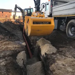 Żółta koparka z chwytakiem umieszcza rurę w wykopie; w tle koparka i ciężarówka z operatorem, budynek gospodarczy w oddali.