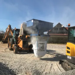 Pomarańczowa koparko-ładowarka na żwirowym terenie napełnia big bag z kruszywem przez metalowy zasobnik; w tle dom jednorodzinny.