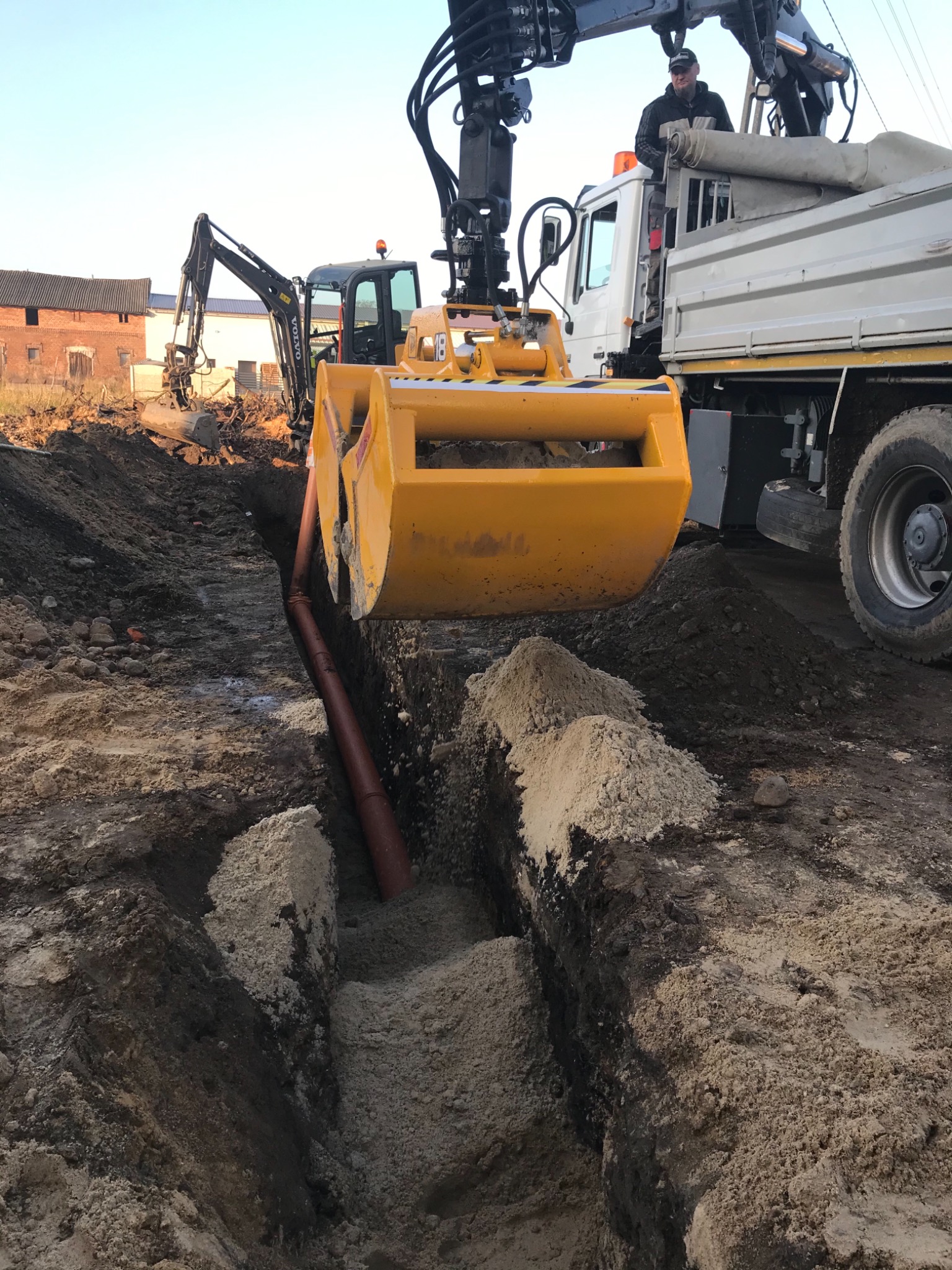 Żółta koparka z chwytakiem umieszcza rurę w wykopie; w tle koparka i ciężarówka z operatorem, budynek gospodarczy w oddali.