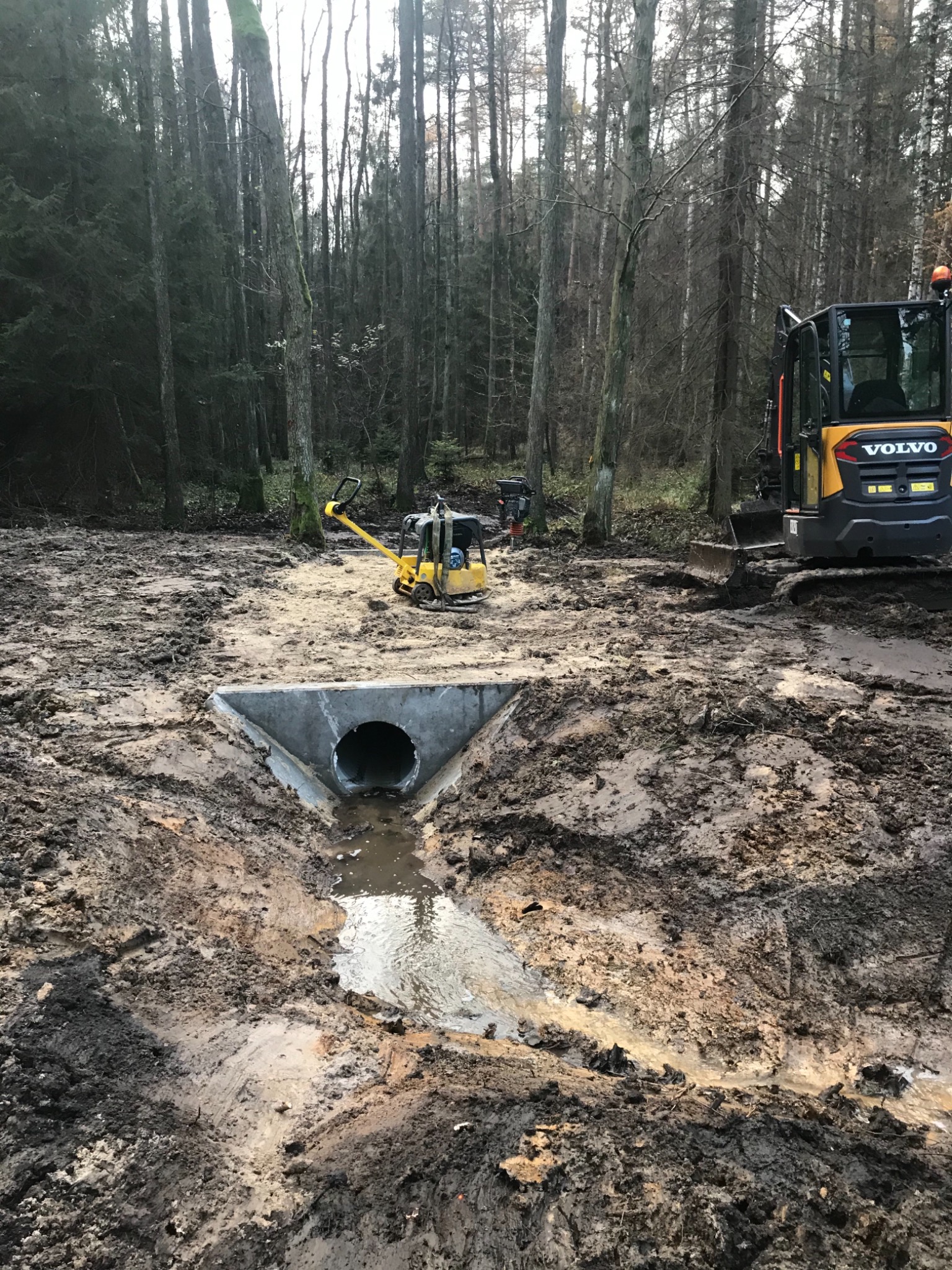 Betonowy przepust rurowy w wykopie, otoczony błotnistym terenem budowy, z widocznymi maszynami budowlanymi w tle, na zalesionym terenie.