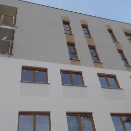 Wielopiętrowy budynek z szarą elewacją górnej części i białą dolnej, z drewnianymi akcentami wokół okien na górze i balkonami w stanie surowym po lewej stronie.