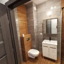 Nowoczesna łazienka z drewnopodobnymi płytkami na podłodze i ścianie, wisząca toaleta z zabudową, biała szafka z umywalką i lustro w czarnej ramie, widok z perspektywy wejścia.