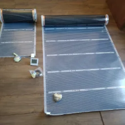 Rozwinięte maty grzewcze podłogowe na drewnianej podłodze, widoczne elementy montażowe i sterownik temperatury.