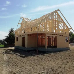 Wznoszenie więźby dachowej z drewna na budynku w stanie surowym otwartym, widoczne płyty OSB na ścianach i ziemia wokół budynku z wyraźnymi śladami opon.