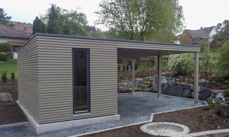 Nowoczesna konstrukcja z drewna i metalu: budynek z oknem obok zadaszonego miejsca postojowego, otoczone kamieniami i roślinami w ogrodzie.