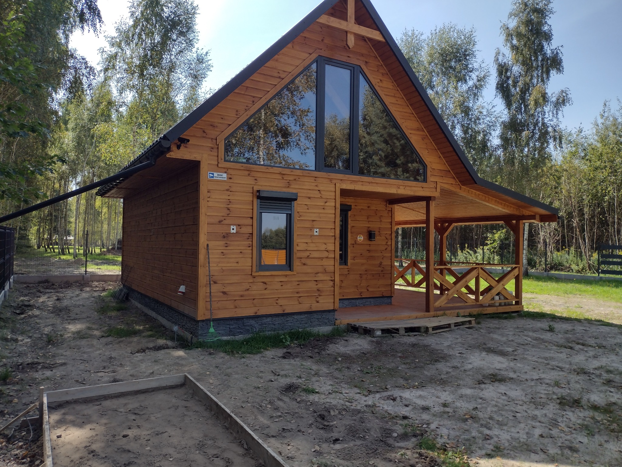 Drewniany dom z dużym trójkątnym oknem i zadaszonym tarasem, widoczne rynny i przygotowany teren pod wylewkę.