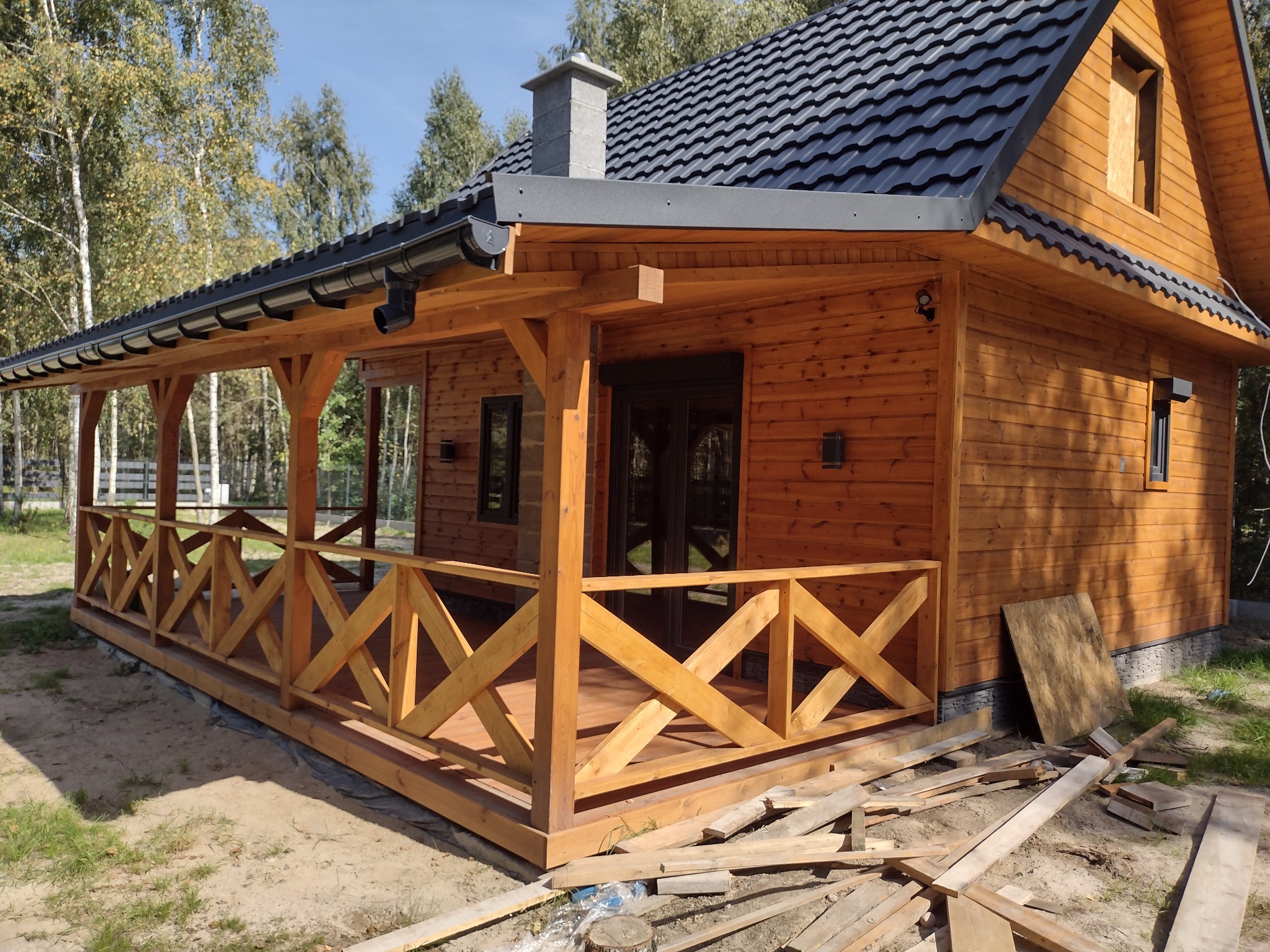 Drewniany dom z zadaszonym tarasem w trakcie budowy, widoczne elementy konstrukcyjne, czarne rynny i fragmenty poszycia dachowego, teren wokół domu częściowo zagospodarowany.