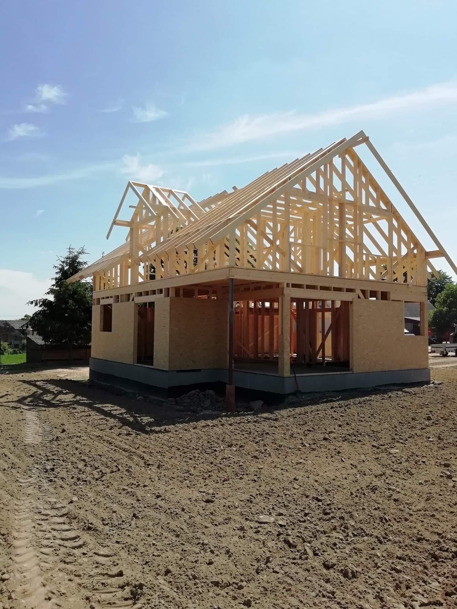 Wznoszenie więźby dachowej z drewna na budynku w stanie surowym otwartym, widoczne płyty OSB na ścianach i ziemia wokół budynku z wyraźnymi śladami opon.