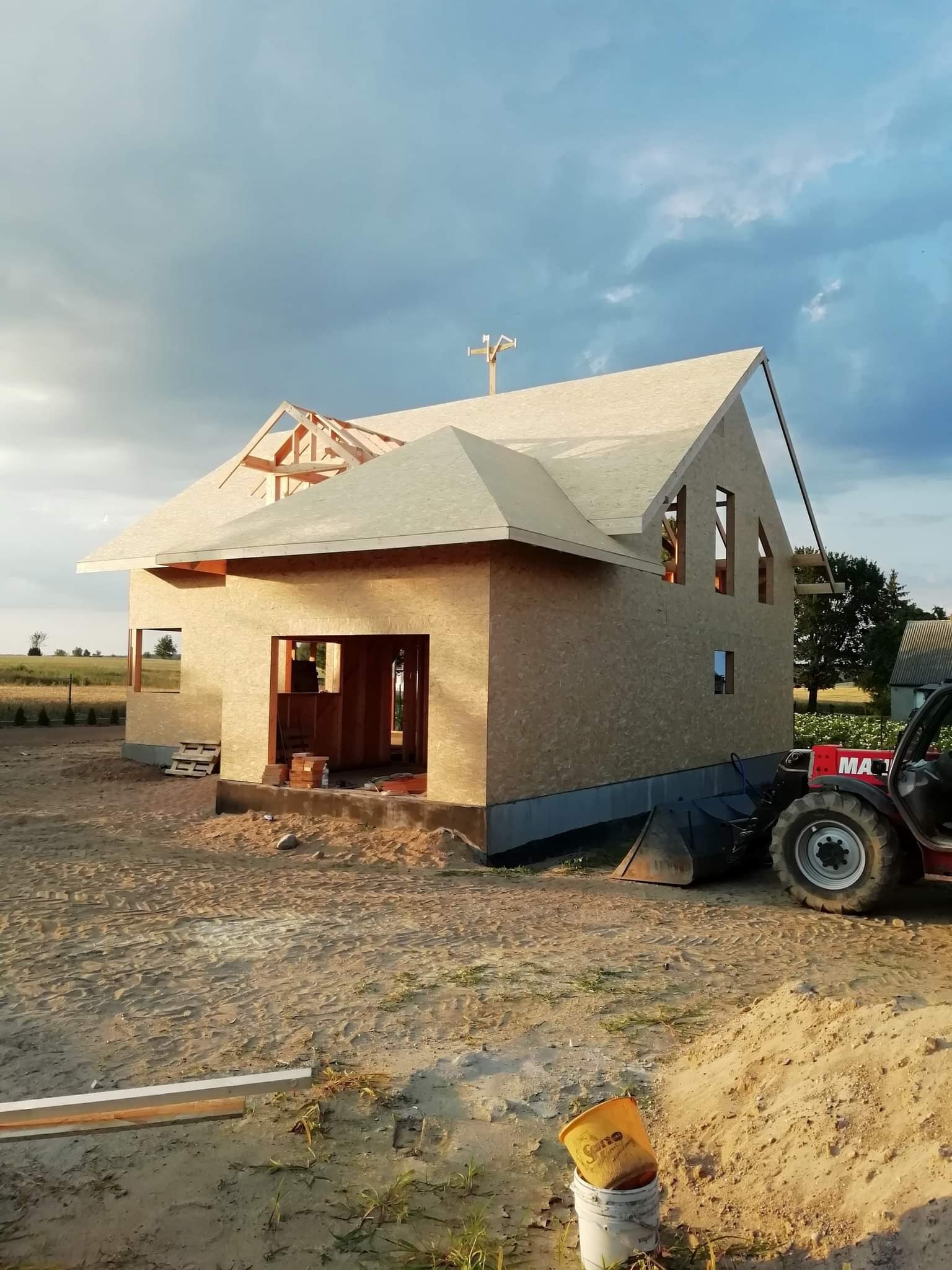Surowy stan domu szkieletowego z widoczną więźbą dachową w budowie, ściany obite płytami OSB, w tle krajobraz polny i maszyna budowlana.