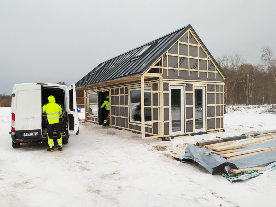 Budowa małego domu szkieletowego w zimowym krajobrazie; widoczna blacha na dachu, drewniana konstrukcja ścian z oknami, pracownicy w odblaskowych ubraniach oraz dostawczy samochód z otwartymi...