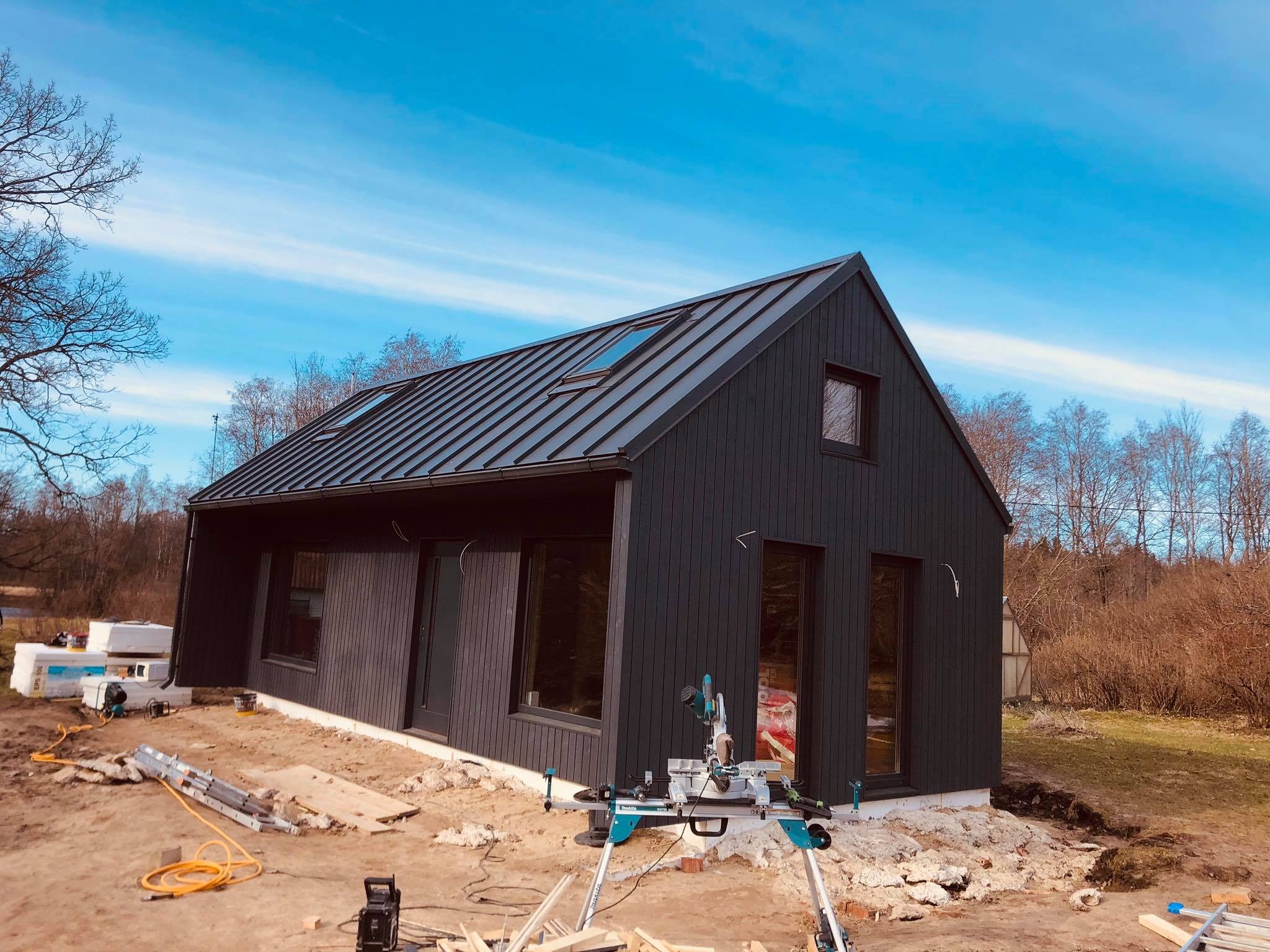 Nowoczesny dom z ciemną, pionową elewacją z desek i blachą trapezową na dachu, widoczna piła ukośnica na placu budowy.