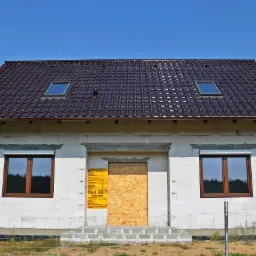 Surowy stan domu jednorodzinnego z brązowym dachem, dwoma oknami dachowymi, oknami z roletami zewnętrznymi i deskami zasłaniającymi wejście.