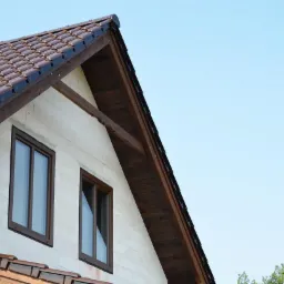 Fragment domu z brązowymi oknami, brązowym dachem z dachówki i białymi ścianami z betonu komórkowego na tle błękitnego nieba.