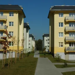 Nowoczesne, pastelowe bloki mieszkalne z balkonami w słoneczny dzień. Widok na osiedle z chodnikiem i młodymi drzewami.