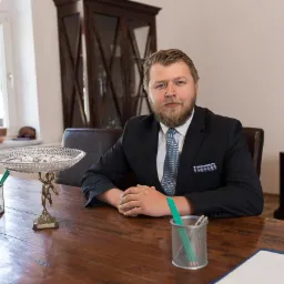 Mężczyzna w garniturze siedzi za biurkiem z drewnianym blatem, na którym stoi kryształowa misa na postumencie w kształcie figurki, metalowy pojemnik z ołówkami i kartki papieru.