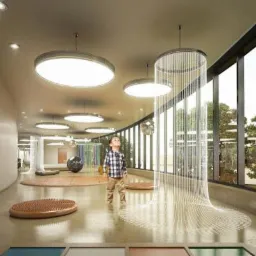 Jasne, przestronne wnętrze przedszkola z okrągłymi lampami sufitowymi, dekoracyjną instalacją z białych włókien i małym chłopcem w koszuli w kratę, spoglądającym w górę.