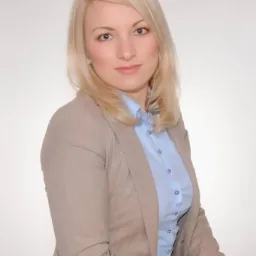 Portret uśmiechniętej blondynki w beżowej marynarce i błękitnej koszuli na jasnym tle.