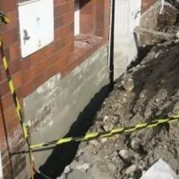 Świeżo zaizolowany fundament budynku z cegły, zabezpieczony żółto-czarną taśmą ostrzegawczą, widoczny wykop z ziemią.