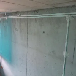 Surowa ściana z betonu w Gdyni z widocznymi, białymi rurkami instalacyjnymi przymocowanymi do ściany i sufitu, zabezpieczonymi gipsem w miejscu łączenia.