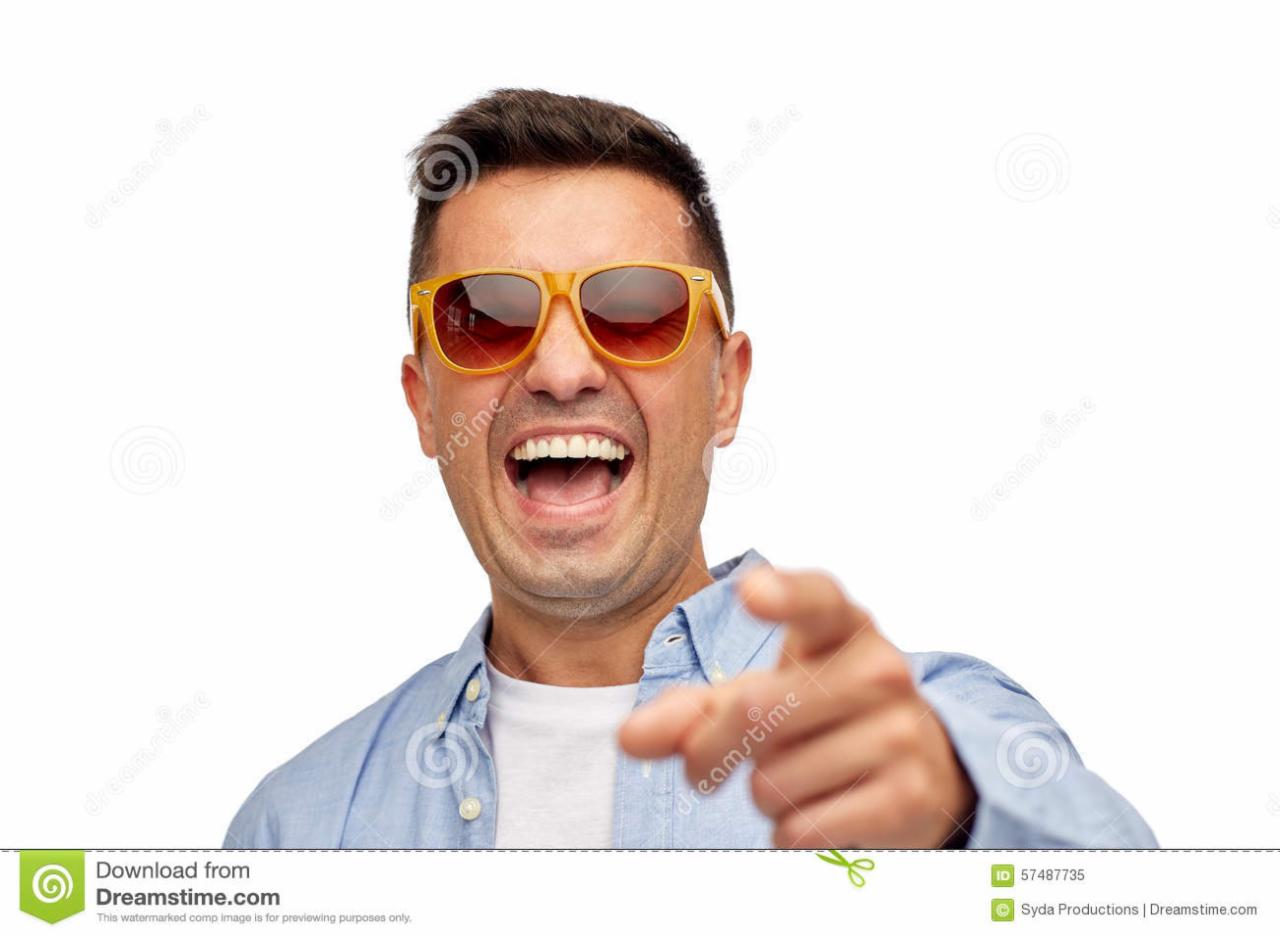 Mężczyzna w pomarańczowych okularach, z otwartymi ustami, wskazuje palcem w stronę widza na białym tle. Ubrany w niebieską koszulę. Uśmiech i ekspresja.