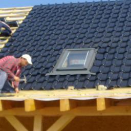 KONCEPT Justyna Łuczak - Montaż czarnej dachówki ceramicznej z oknem dachowym; widoczni dekarze w trakcie pracy, drewniana konstrukcja dachu.