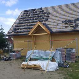 KONCEPT Justyna Łuczak - Budowa domu w trakcie: widok na częściowo pokryty dach z czarną dachówką, zabezpieczony membraną, z widoczną konstrukcją drewnianą, rusztowaniami i materiałami budowlanymi na ziemi, w tle drzewa...