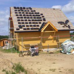 KONCEPT Justyna Łuczak - Wznoszenie domu w technologii szkieletowej: montaż dachu z czarnej dachówki ceramicznej, widoczne rusztowania i materiały budowlane na placu budowy.