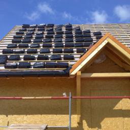 KONCEPT Justyna Łuczak - Fragment dachu w trakcie krycia dachówką ceramiczną, widoczny drewniany szkielet konstrukcji, membrany i łaty, z bliska ułożone rzędy dachówek na połaci dachu pod błękitnym niebem.