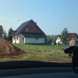 KONCEPT Justyna Łuczak - Widok zza szyby samochodu na plac budowy z kilkoma domami w tle, jeden z nich w trakcie budowy z widocznym stosem ziemi.