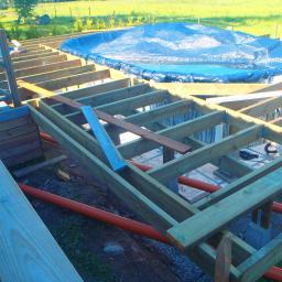 KONCEPT Justyna Łuczak - Konstrukcja drewniana tarasu w trakcie budowy, widoczne legary, słupy betonowe, rury kanalizacyjne i zadaszony basen w tle.