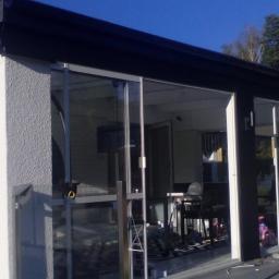 KONCEPT Justyna Łuczak - Nowoczesna przeszklona weranda z przesuwnymi drzwiami i widocznym wnętrzem z meblami ogrodowymi, zewnętrzna elewacja wykończona tynkiem strukturalnym.