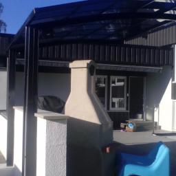 KONCEPT Justyna Łuczak - Zadaszony taras z kominkiem zewnętrznym, widok na dom z czarną elewacją i model żaglówki w oknie.