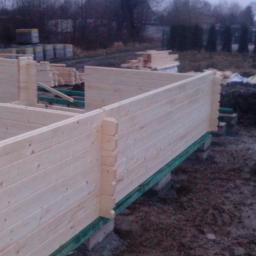 KONCEPT Justyna Łuczak - Wznoszenie ścian z drewnianych bali na fundamencie, widoczne połączenia bali w narożnikach, w tle stosy materiałów budowlanych i drzewa.
