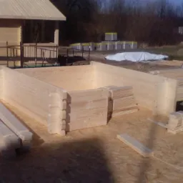 Wznoszenie konstrukcji z drewnianych bali: widoczne ściany w trakcie budowy, przygotowane elementy i narzędzia na placu budowy, wstępny etap montażu.