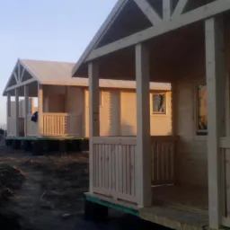 Dwa surowe domki letniskowe z drewna w trakcie budowy, widoczne rusztowania w tle, teren nierówny, wczesny etap prac.