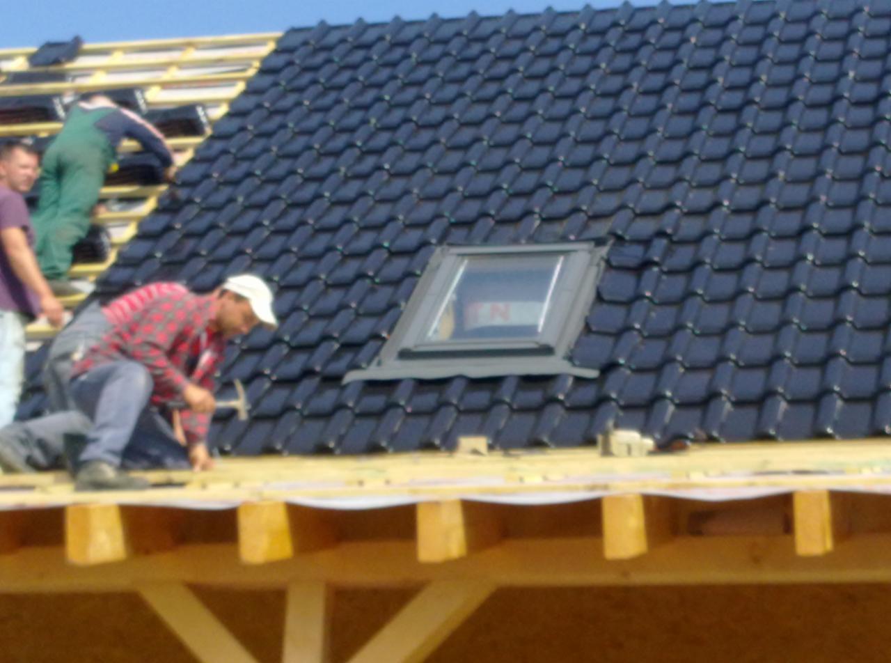 Montaż czarnej dachówki ceramicznej z oknem dachowym; widoczni dekarze w trakcie pracy, drewniana konstrukcja dachu.