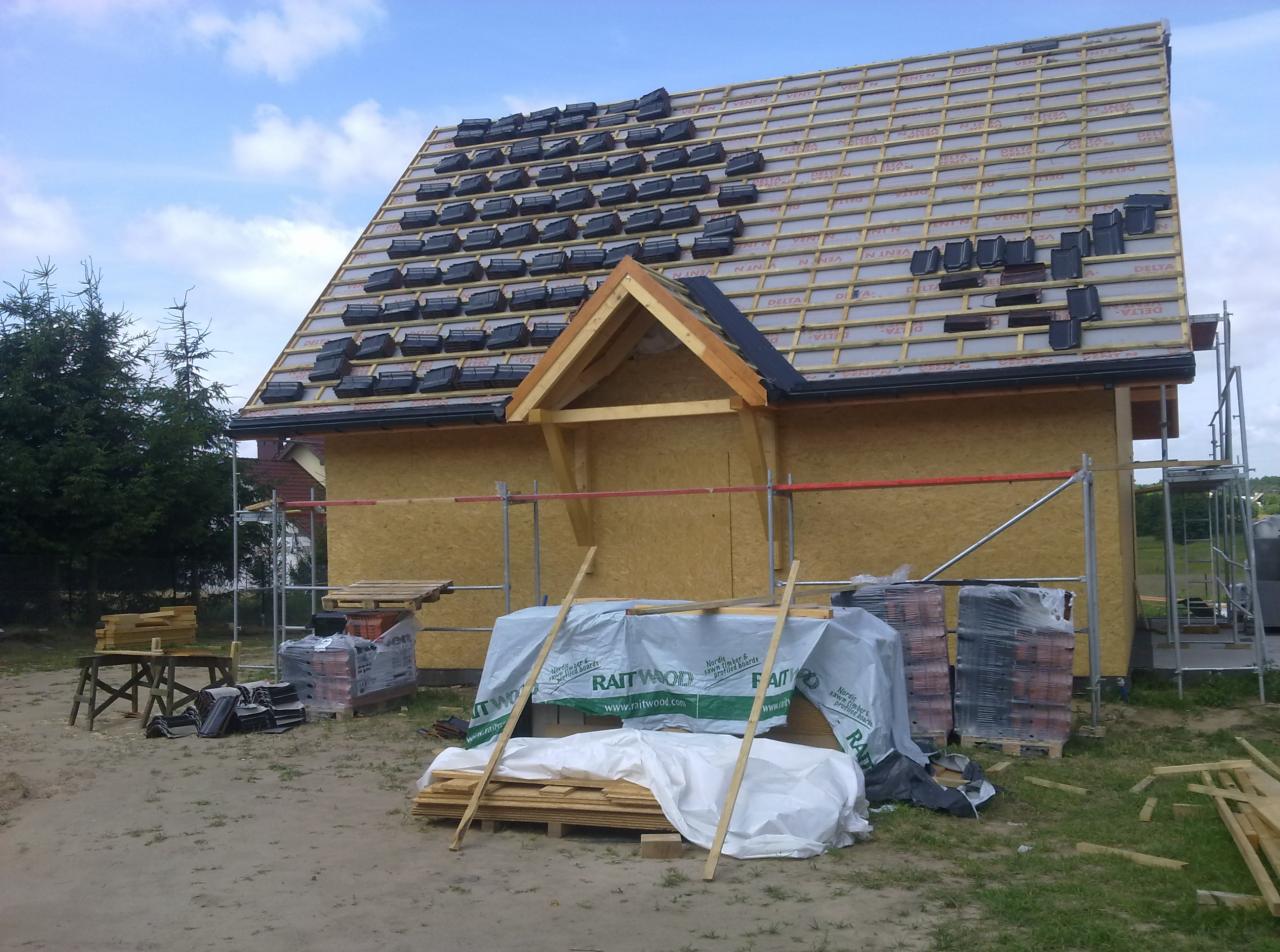 Budowa domu w trakcie: widok na częściowo pokryty dach z czarną dachówką, zabezpieczony membraną, z widoczną konstrukcją drewnianą, rusztowaniami i materiałami budowlanymi na ziemi, w tle drzewa...